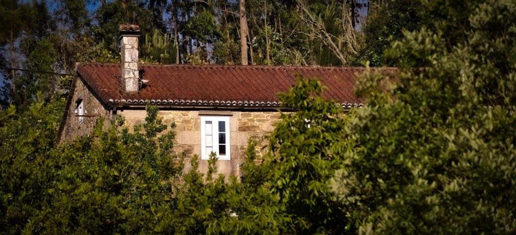 Casa antiga de granito com telhado de telha vermelha, chaminé de pedra e uma janela branca, meio escondida pelas árvores