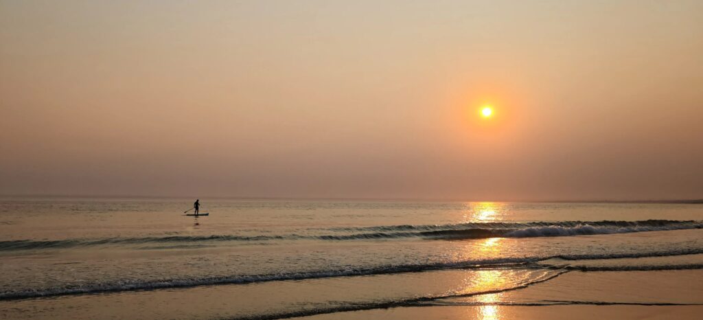 Pequeña silueta de una persona haciendo paddle surf sobre agua en calma, con el sol reflejado en el mar