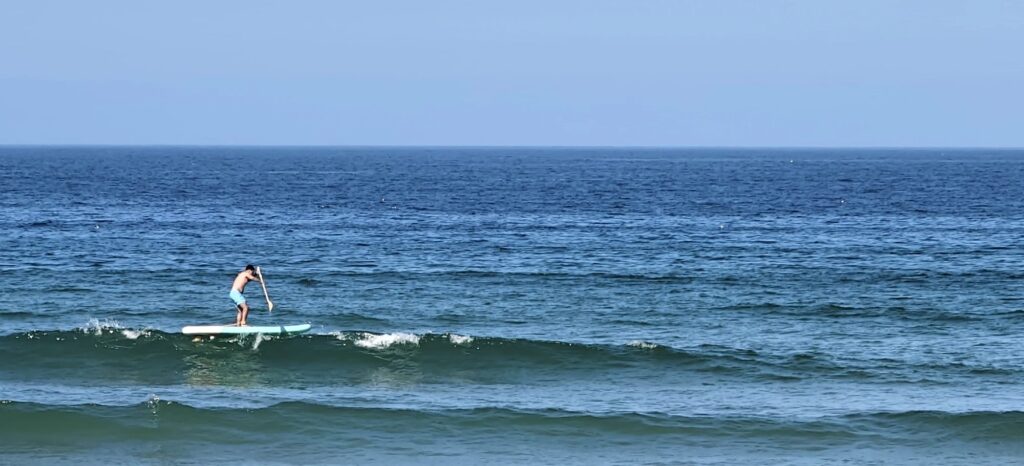 Persoa facendo paddle surf sobre unha onda pequena nun mar azul e despexado na Praia da Rainha
