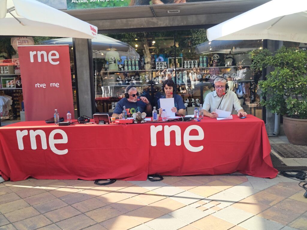 Mesa de la emisión en directo de RNE en el Zoo Aquarium de Madrid, con tres locutores con auriculares y un pequeño peluche de elefante sobre el mantel rojo de RNE