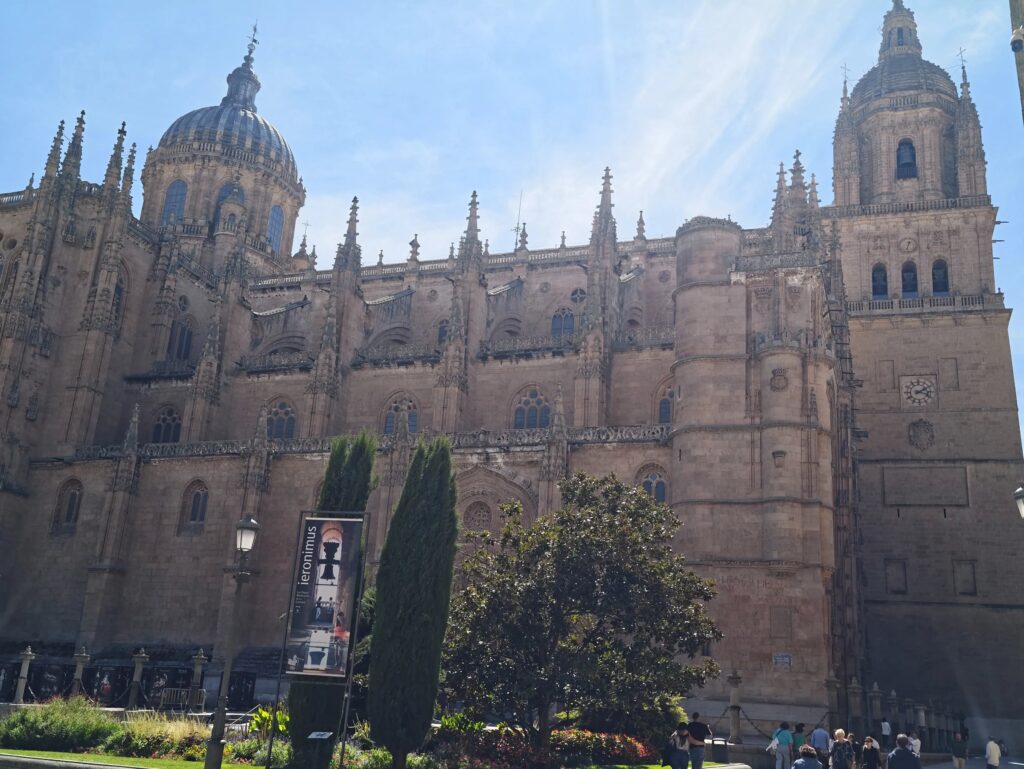 Lateral de la Catedral Nueva con la cúpula y la torre sobre cipreses y un jardín, con rayos de sol en el cielo