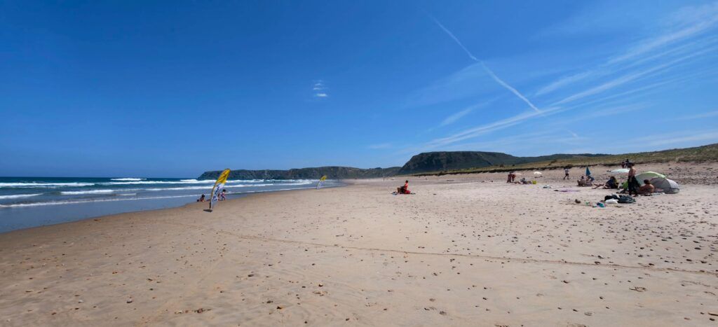 Amplia playa de arena en Xagó con banderas amarillas de escuela de surf, dunas bajas y un cabo de cima plana al fondo bajo un cielo azul