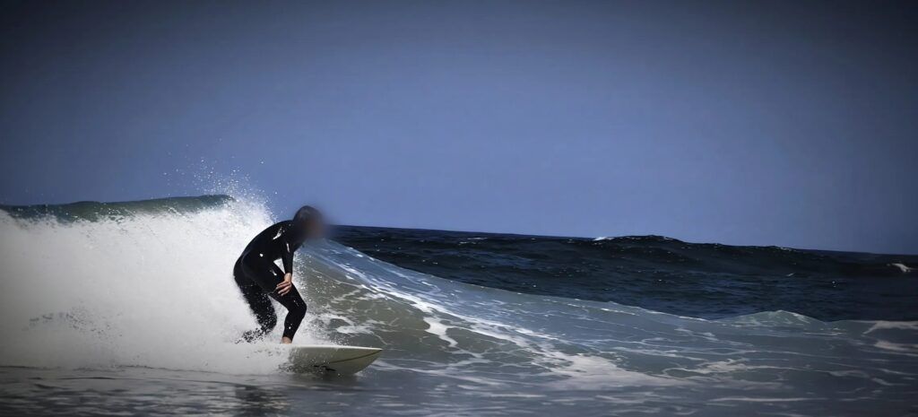 Surfista con neopreno negro agachado sobre una tabla blanca mientras la ola rompe detrás