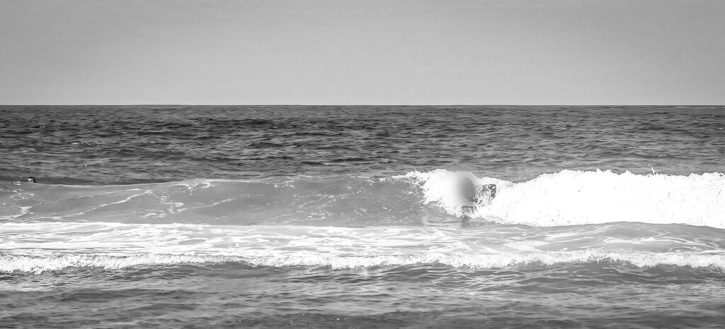 Fotografía en blanco y negro de una surfista cogiendo una ola pequeña con un bodyboard, con el horizonte plano al fondo
