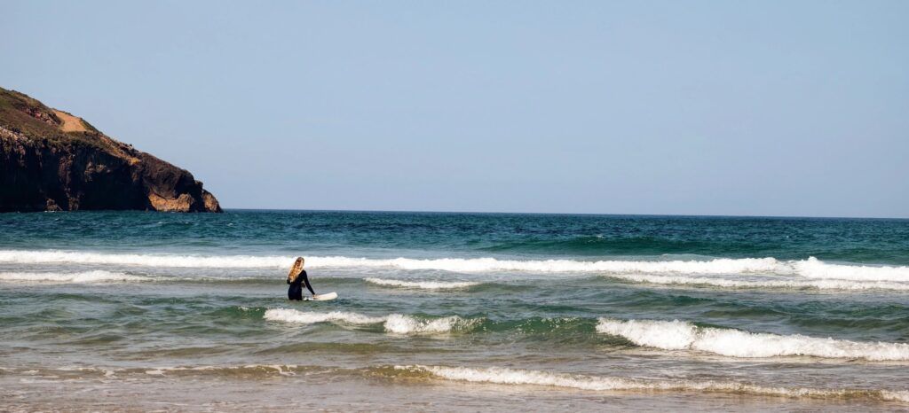 Vista amplia del mar con una surfista con el agua por la cintura sujetando la tabla, y el cabo y los acantilados a la izquierda