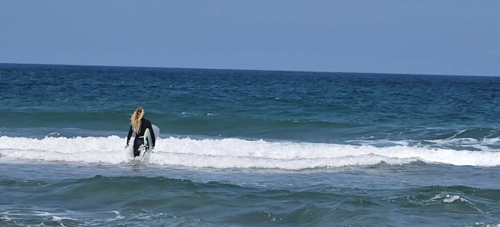 Surfista con traje de neopreno negro y melena clara entrando en la espuma con una tabla blanca