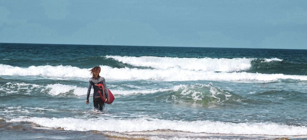 Surfista con neopreno gris y negro y pelo rizado entrando al agua con una tabla roja entre filas de olas rompiendo