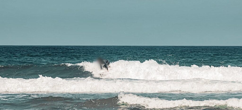 Surfista agachada sobre la tabla dentro de la espuma, con el mar en tonos turquesa y el cielo pálido