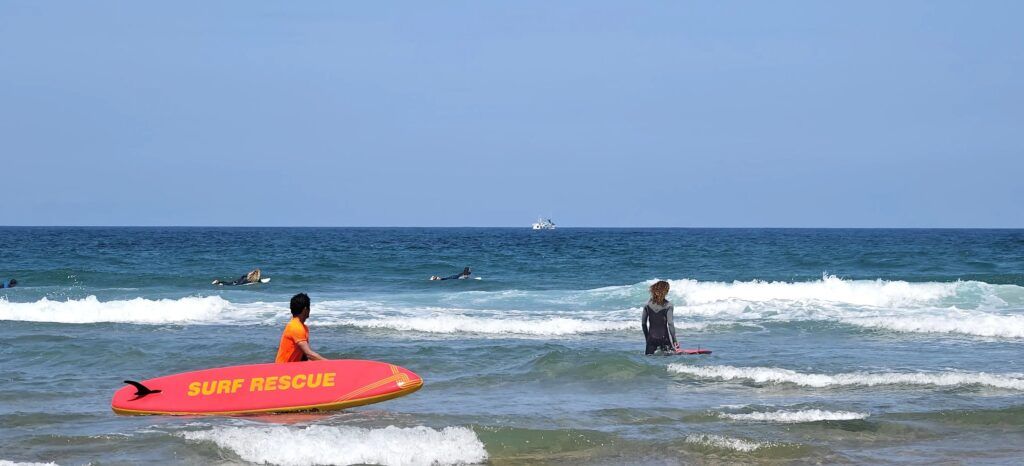 Socorrista con camiseta naranja entrando al agua con una tabla de rescate roja mientras los surfistas esperan más allá de la rompiente