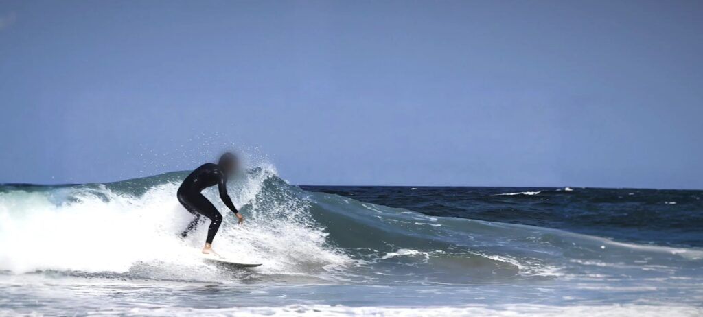 Surfista deslizándose con una tabla blanca por la pared de una ola que rompe, con la espuma saliendo por detrás