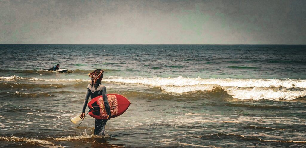 Fotografía en tonos cálidos de una surfista con tabla roja entrando al agua mientras otro rema más allá de la orilla